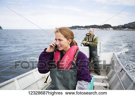 Stock Photograph - Set-netter talking on a cell phone while her husband drives a set-net skiff in Seldovia Bay, South-central Alaska; Alaska, United States of America. Fotosearch