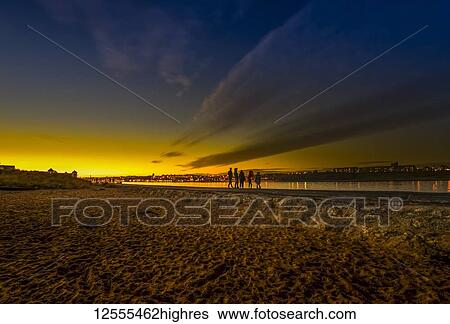 Stock Photograph - Silhouette of a family with three children standing at the water's edge of the River Tyne at sunset and a beach in the foreground; South Shields, Tyne and Wear, England. Fotosearch