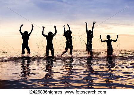 Stock Photograph - Silhouette of five children jumping out of the water in a row at sunset, Sandbanks Provincial Park; Picton, Ontario, Canada. Fotosearch