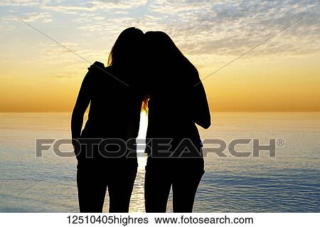 Stock Photograph - Silhouette of two teenage girls standing in an embrace looking out at a lake at sunset, Woodbine Beach; Toronto, Ontario, Canada. Fotosearch