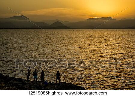 Stock Photograph - Silhouettes of fishermen at sunset; Alcudia, Mallorca, Balearic Islands, Spain. Fotosearch