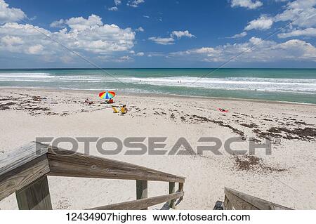Sunbathers sit on the sand of Pelican Beach with a colourful umbrella looking out to the Atlantic ocean, and wooden steps with railing in the foreground; Satellite Beach, Florida, United States of America View Large Photo Image Stock Photograph - Sunbathers sit on the sand of Pelican Beach with a colourful umbrella looking out to the Atlantic ocean, and wooden steps with railing in the foreground; Satellite Beach, Florida, United States of America. Fotosearch
