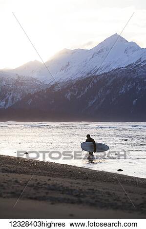 Stock Photograph - Surfer Carries His Board Out Of Kachemak Bay, Homer Spit, Southcentral Alaska, USA. Fotosearch