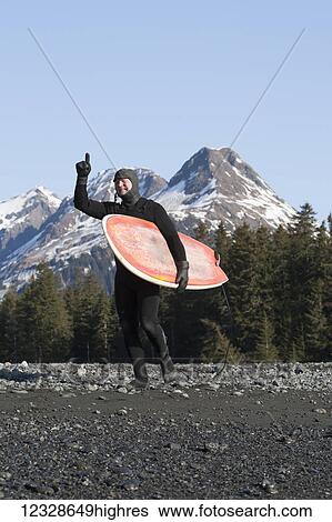 Surfer carrying his surfboard and pointing his finger upwards along the shore of the Kenai Peninsula Outer Coast, South-central Alaska; Alaska, United States of America View Large Photo Image Stock Photograph - Surfer carrying his surfboard and pointing his finger upwards along the shore of the Kenai Peninsula Outer Coast, South-central Alaska; Alaska, United States of America. Fotosearch