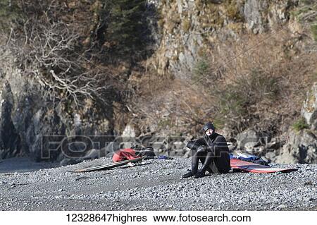 Stock Photograph - Surfer hanging out on the beach with his surfboard, Kenai Peninsula Outer Coast, South-central Alaska; Alaska, United States of America. Fotosearch
