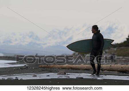 Stock Photograph - Surfer Holding His Surfboard On The Shore Near Yakutat, Southeast Alaska, USA. Fotosearch