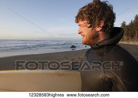 Stock Photograph - Surfer holding surfboard looking at ocean at sunset, Southeast Alaska; Yakutat, Alaska, United States of America. Fotosearch