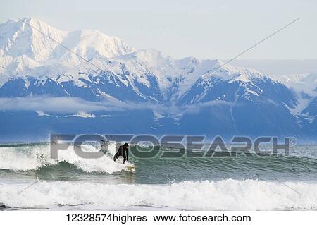 Stock Photograph - Surfer riding a wave with Fairweather Range in the background, Southeast Alaska; Yakutat, Alaska, United States of America. Fotosearch