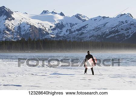 Stock Photograph - Surfer standing in the ocean along the Kenai Peninsula Outer Coast, South-central Alaska; Alaska, United States of America. Fotosearch