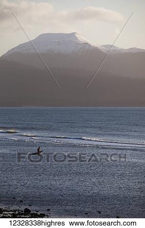 Stock Photograph - Surfer Standing In The Waters Of Kachemak Bay At Sunset With Kenai Mountains In The Background, Homer Spit, Southcentral Alaska, USA. Fotosearch