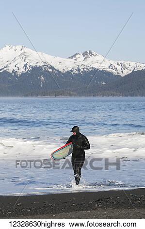 Stock Photograph - Surfer walking in the water on the Kenai Peninsula Outer Coast, South-central Alaska; Alaska, United States of America. Fotosearch