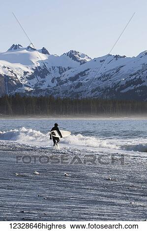 Stock Photograph - Surfer walking into the ocean along the Kenai Peninsula Outer Coast, South-central Alaska; Alaska, United States of America. Fotosearch