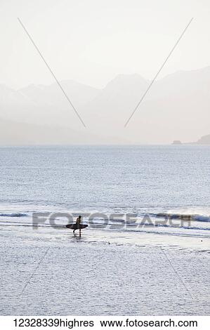 Stock Photograph - Surfer Walking On Beach, Kachemak Bay With Kenai Mountains In The Background, Homer Spit, Southcentral Alaska, USA. Fotosearch
