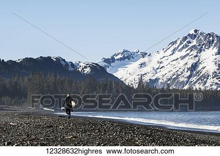 Stock Photograph - Surfer walking on the beach along the Kenai Peninsula Outer Coast, South-central Alaska, USA. Fotosearch