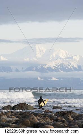 Stock Photograph - Surfer with board entering the ocean with Fairweather Range in the distance, Southeast Alaska; Yakutat, Alaska, United States of America. Fotosearch