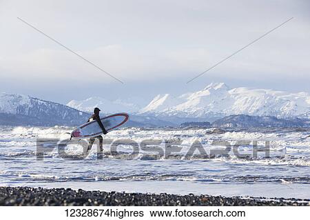 Stock Photograph - Surfer with surfboard entering Kachemak Bay, South-central Alaska; Homer, Alaska, United States of America. Fotosearch