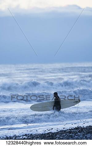 Stock Photograph - Surfer with surfboard standing in the splashing water along the shore, Kachemak Bay, South-central Alaska; Homer, Alaska, United States of America. Fotosearch