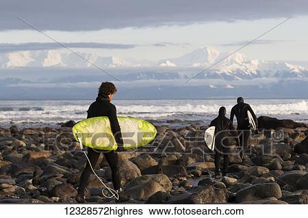 Stock Photograph - Surfers on rocky beach with Fairweather Range in the background, Southeast Alaska; Yakutat, Alaska, United States of America. Fotosearch