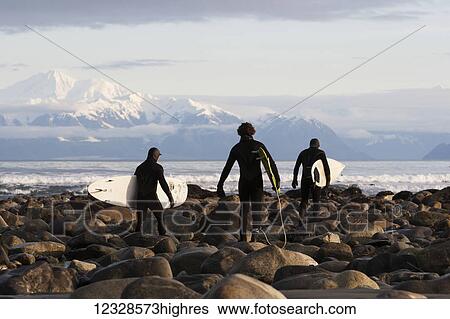 Stock Photograph - Surfers on rocky beach with Fairweather Range in the background, Southeast Alaska; Yakutat, Alaska, United States of America. Fotosearch