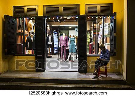 Stock Photograph - Tailor shop at night with a man sitting on a stool outside the door; Hoi An, Quang Nam, Vietnam. Fotosearch
