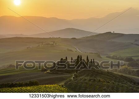Stock Photograph - The morning sun rising above layered Tuscany green hills and the small Podere Belvedere villa with a vineyard in the foreground near San Quirico D'orcia; Tuscany, Italy. Fotosearch