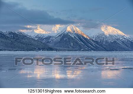 Stock Photograph - The warm light of sunset lights up the snow-covered peaks of Turnagain Pass, the mountains reflecting on the icy ocean surface, Seward Highway, South-central Alaska in winter; Alaska, United States of America. Fotosearch