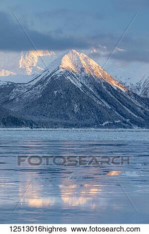 Stock Photograph - The warm light of sunset lights up the snow-covered peaks of Turnagain Pass, the mountains reflecting on the icy ocean surface, Seward Highway, South-central Alaska in winter; Alaska, United States of America. Fotosearch
