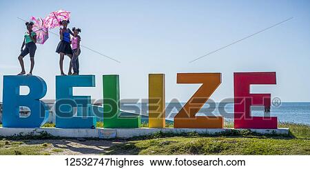 Stock Photograph - Three girls stand on the colourful Belize sign with a view of the ocean behind; Belize City, Belize. Fotosearch