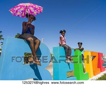 Stock Photograph - Three girls stand on the colourful Belize sign; Belize City, Belize. Fotosearch