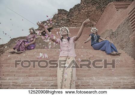Stock Photograph - Three women in traditional dress portraying happiness as they throw flower petals and smile; Bagan, Myanmar. Fotosearch