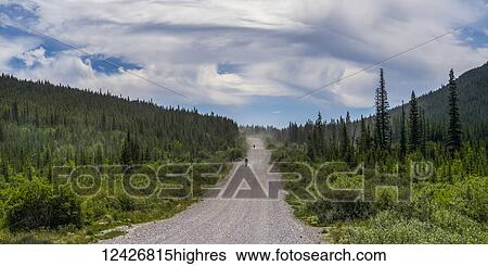 Stock Photograph - Two cyclists ride down a gravel road between hills and forest; Canada. Fotosearch