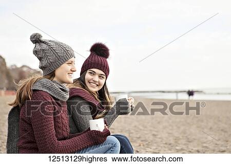 Stock Photograph - Two friends at the beach wearing knit hats and scarves, sitting and having coffee together, Woodbine Beach; Toronto, Ontario, Canada. Fotosearch