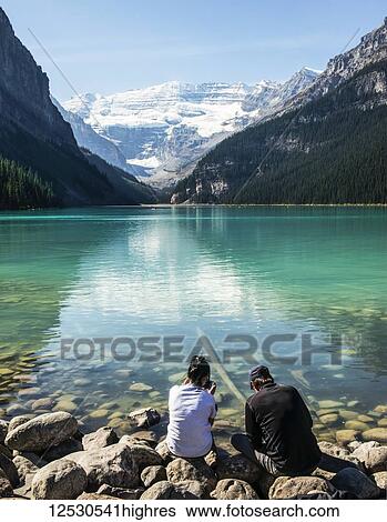 Stock Photograph - Two people distracted by looking at their smart phones while sitting along the shoreline of Lake Louise; Alberta Canada. Fotosearch