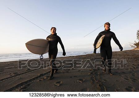 Stock Photograph - Two surfers walking on a beach with their boards, Southeast Alaska; Yakutat, Alaska, United States of America. Fotosearch