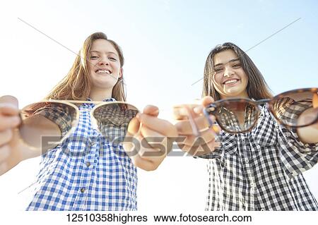 Stock Photograph - Two teenage girls posing and holding out their sunglasses towards the camera as if offering them to the viewer, Woodbine Beach; Toronto, Ontario, Canada. Fotosearch
