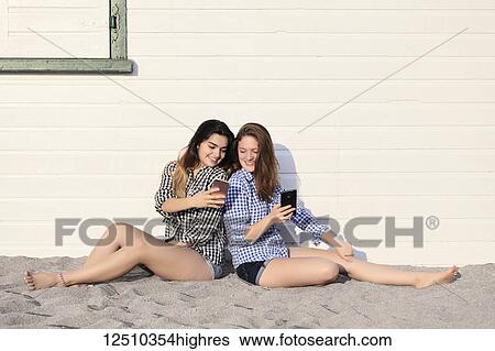 Two teenage girls sitting in the sand against a building looking at their smart phones together, Woodbine Beach; Toronto, Ontario, Canada View Large Photo Image Stock Photograph - Two teenage girls sitting in the sand against a building looking at their smart phones together, Woodbine Beach; Toronto, Ontario, Canada. Fotosearch