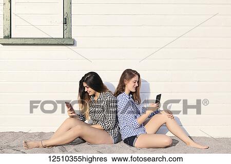 Stock Photograph - Two teenage girls sitting in the sand against a building looking at their smart phones together, Woodbine Beach; Toronto, Ontario, Canada. Fotosearch