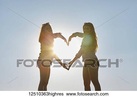 Stock Photograph - Two teenage girls stand making the shape of a heart with their arms as the sunlight shines through, Woodbine Beach; Toronto, Ontario, Canada. Fotosearch