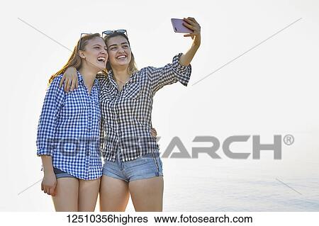Two teenage girls take a self-portrait as they stand in an embrace at sunset at the lake, Woodbine Beach; Toronto, Ontario, Canada View Large Photo Image Stock Photograph - Two teenage girls take a self-portrait as they stand in an embrace at sunset at the lake, Woodbine Beach; Toronto, Ontario, Canada. Fotosearch