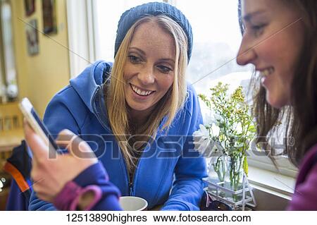 Stock Photograph - Two young women on a smart phone and enjoying coffee at a cafe in downtown Anchorage, South-central Alaska; Anchorage, Alaska, United States of America. Fotosearch