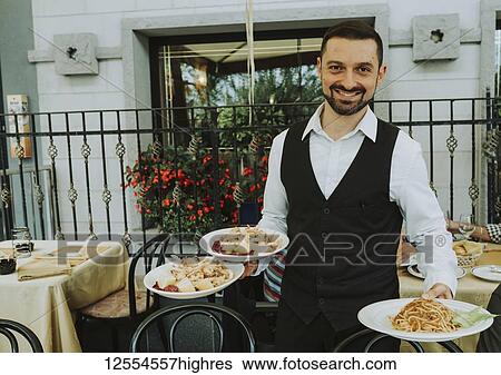 Stock Photograph - Uniformed waiter serving plates of food and smiling for the camera; Italy. Fotosearch