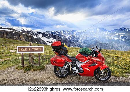 Stock Photograph - View from the Beartooth Highway of the Beartooth Mountains and the sun rays breaking through the clouds. A motorcycle sits on the roadside beside a sign for Bears Tooth; Cody, Wyoming, United States of America. Fotosearch
