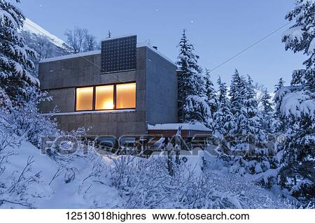 Stock Photograph - Warm light emanates from a cabin in a snow-covered landscape, solar panels on the roof of the cabin, cool light at dusk casts soft light on distant snow-covered Kenai Mountains, South-central Alaska in winter; Moose Pass, Alaska, United States of America. Fotosearch