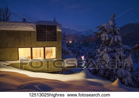 Stock Photograph - Warm light emanates from a cabin in a snow-covered landscape, solar panels on the roof of the cabin, cool light at dusk casts soft light on distant snow-covered Kenai Mountains, South-central Alaska in winter; Moose Pass, Alaska, United States of America. Fotosearch