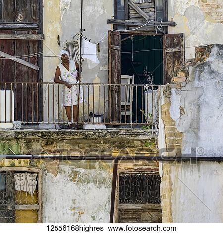 Stock Photograph - Woman stands leaning on the railing of her balcony looking down to the streets below; Havana, Cuba. Fotosearch