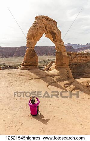 Stock Photograph - Woman taking a cell phone photo of Delicate Arch, Arches National Park; Moab, Utah, United States of America. Fotosearch