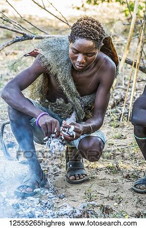 Stock Photograph - Young Hadzabe hunter wearing baboon skin cloak plucks bird killed in morning hunt and prepares to roast it over an open fire near Lake Eyasi; Tanzania. Fotosearch