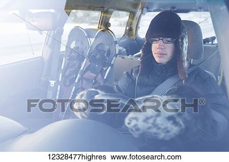 Stock Photograph - Young man driving a car wearing fur hat and mitts with snowshoes in the passenger seat, South-central Alaska; Alaska, United States of America. Fotosearch