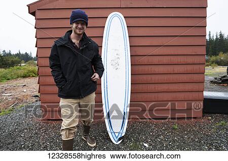 Stock Photograph - Young man standing next to a surfboard, South-central Alaska; Homer, Alaska, United States of America. Fotosearch