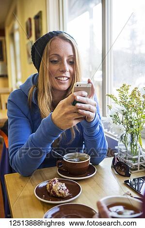 Stock Photograph - Young woman enjoying a cup of tea and pastry while texting on her smart phone in a cafe, South-central Alaska; Anchorage, Alaska, United States of America. Fotosearch